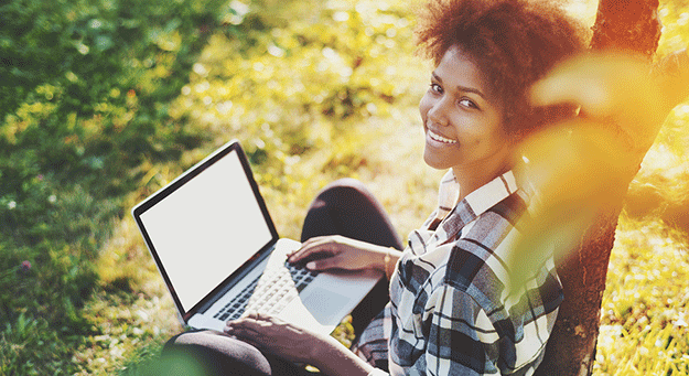 a female sitting outside leaning against a tree and doing research on her laptop