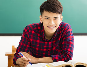 smiling male student sitting at a desk holding a pen and with and open book in front of him