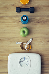 A scale, measuring tape, apple, sports bottle, weight, and orange placed linear on the floor