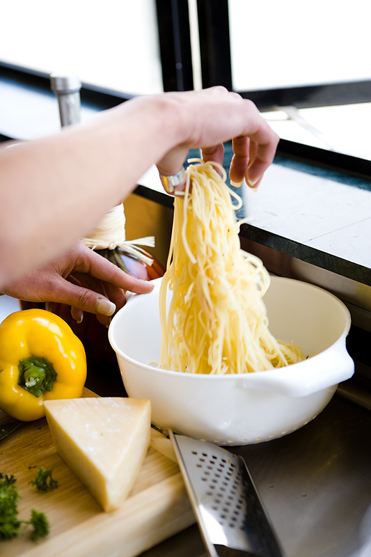 person's hand grabbing spaghetti out of a bowl with a yellow pepper and parmesean cheese on a cutting board to the left of the bowl